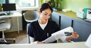 Woman in medical scrubs sits at a desk, looking at papers. She is in a bright, modern office with a chair, desk, plants and window in the background. She appears focused and professional.