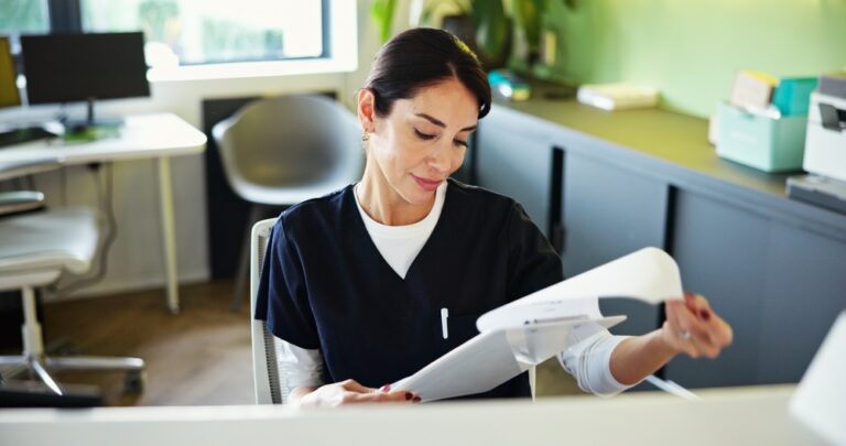 Woman in medical scrubs sits at a desk, looking at papers. She is in a bright, modern office with a chair, desk, plants and window in the background. She appears focused and professional.