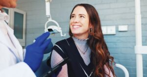 A woman with long brown hair smiles in a dental chair, wearing a navy vest and sheer blouse. A dentist in white coat and blue gloves talks to her, holding dental instruments in a modern dental office.